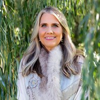 A glamourous older woman stands in front of a willow tree, she is smiling brightly at the camera