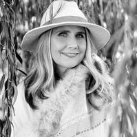 A blonde woman wearing a hat is framed by the branches of a willow tree