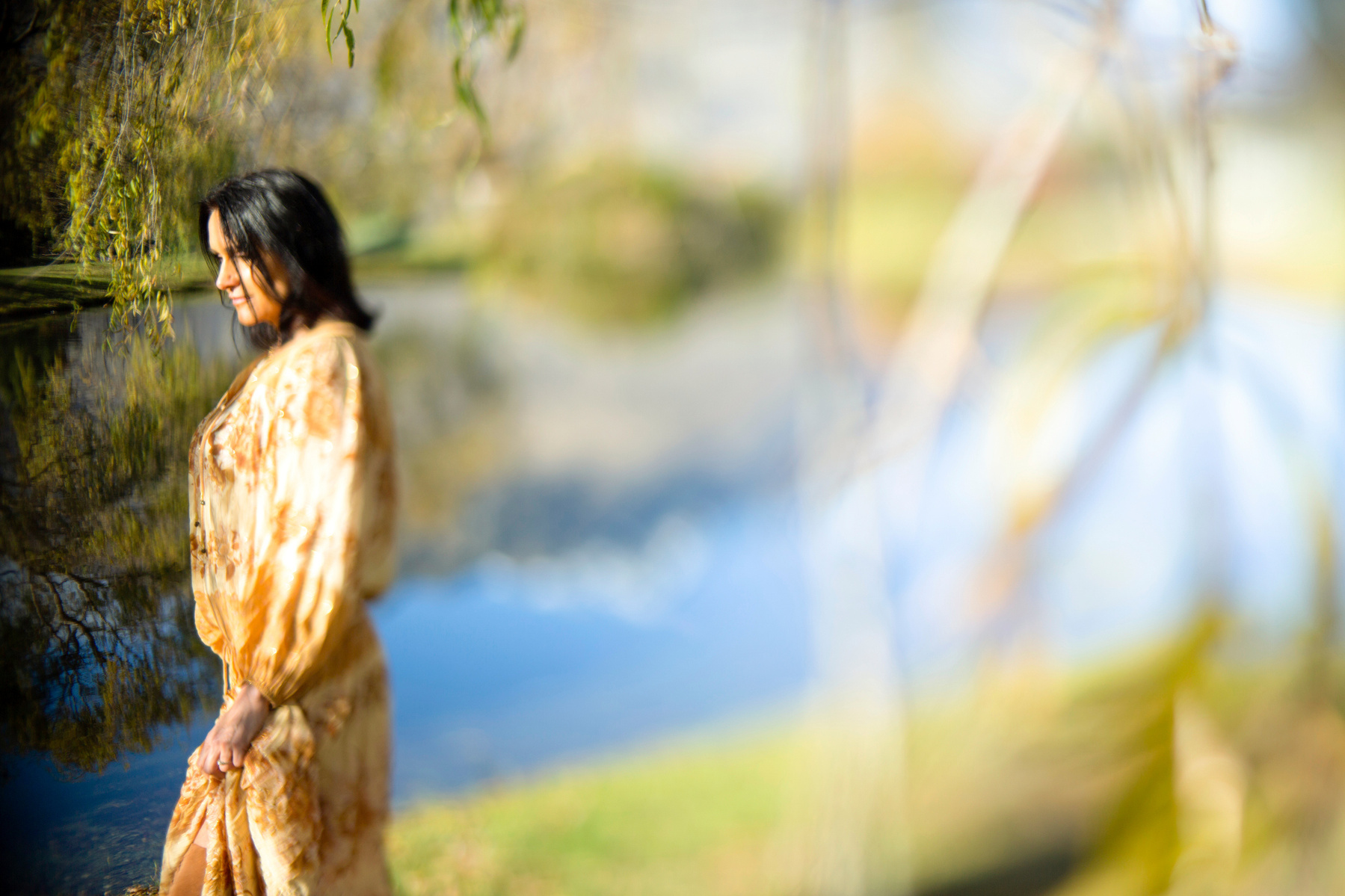 A brunette woman stands by a lake, she looks off camera and the lake is reflecting the mountains behind her