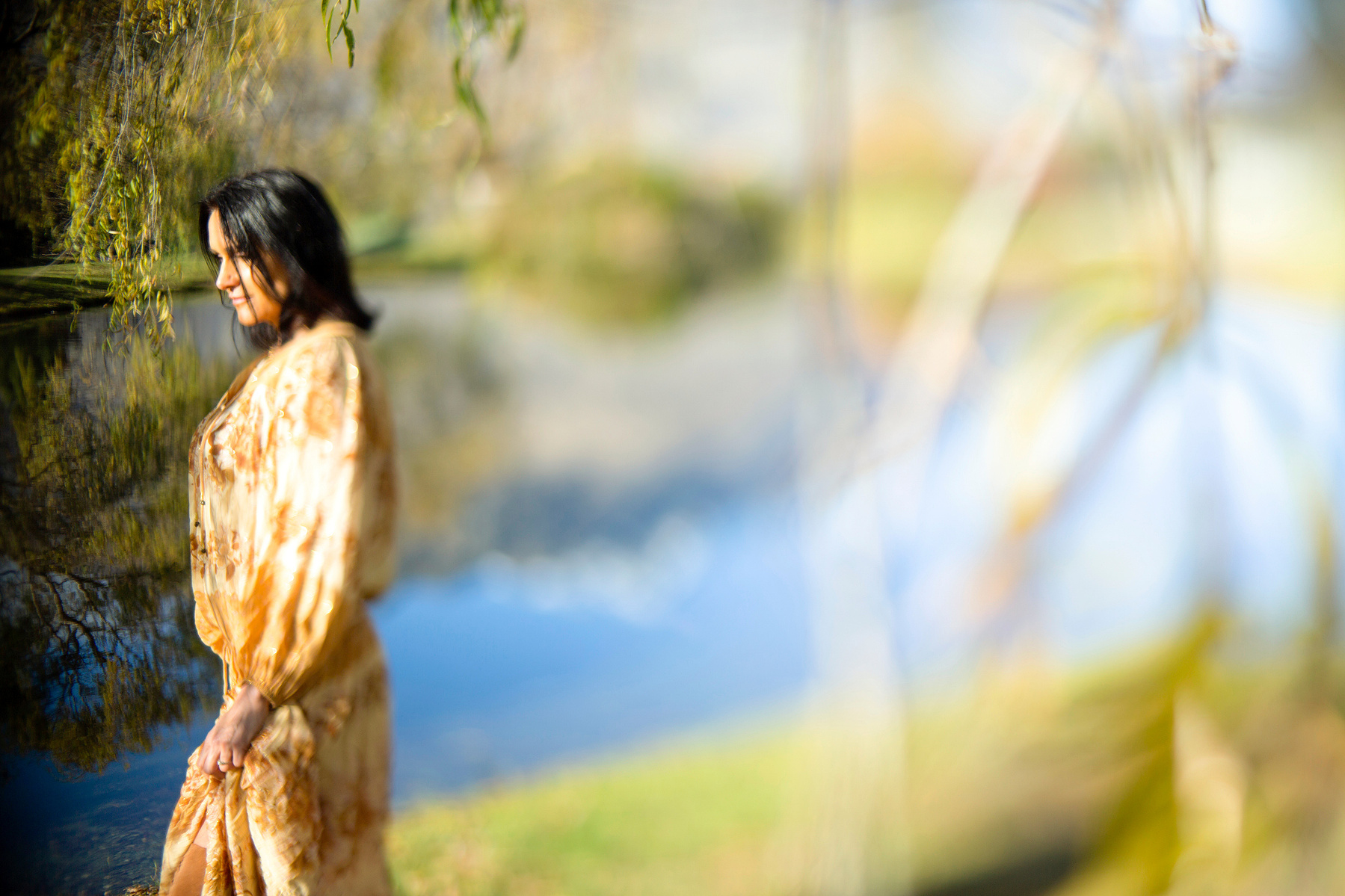 A brunette woman stands by a lake, she looks off camera and the lake is reflecting the mountains behind her