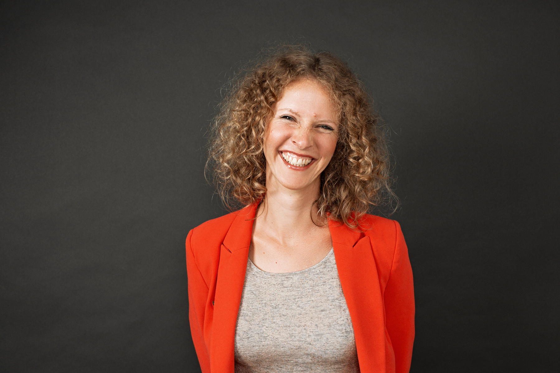 A slim woman with curly hair wears an orange blazer and smiles broadly into the camera