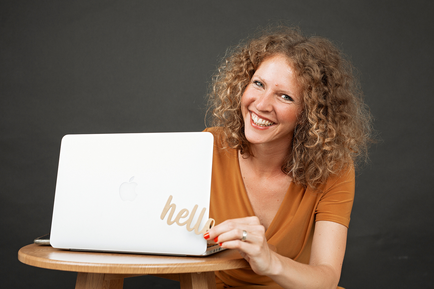 A woman wearing an orange top sits behind a wooden table with a laptiop on, she is holding a sign that spells 'hello' and is smiling