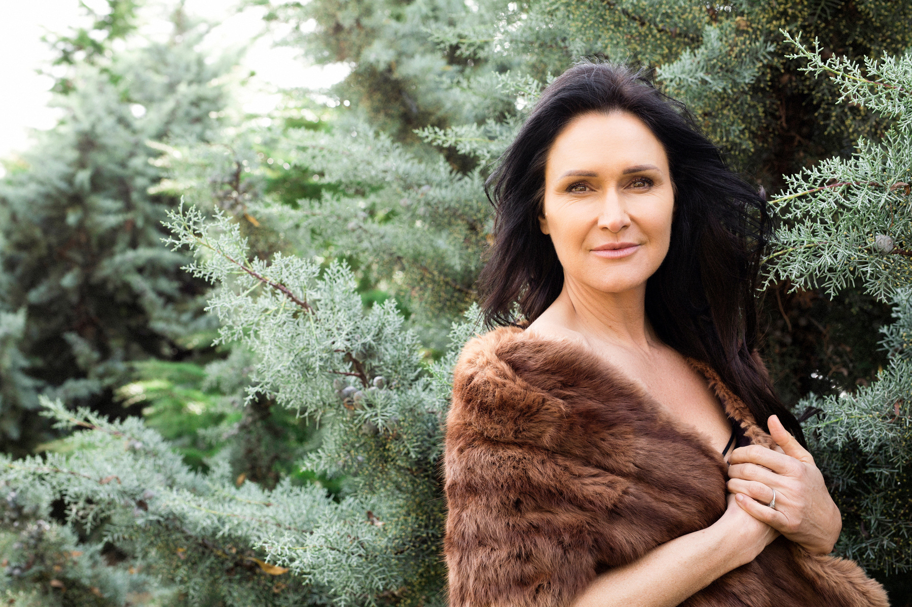 A brunette woman stands in front of frosty pines, she is wrapped in a fur jacket