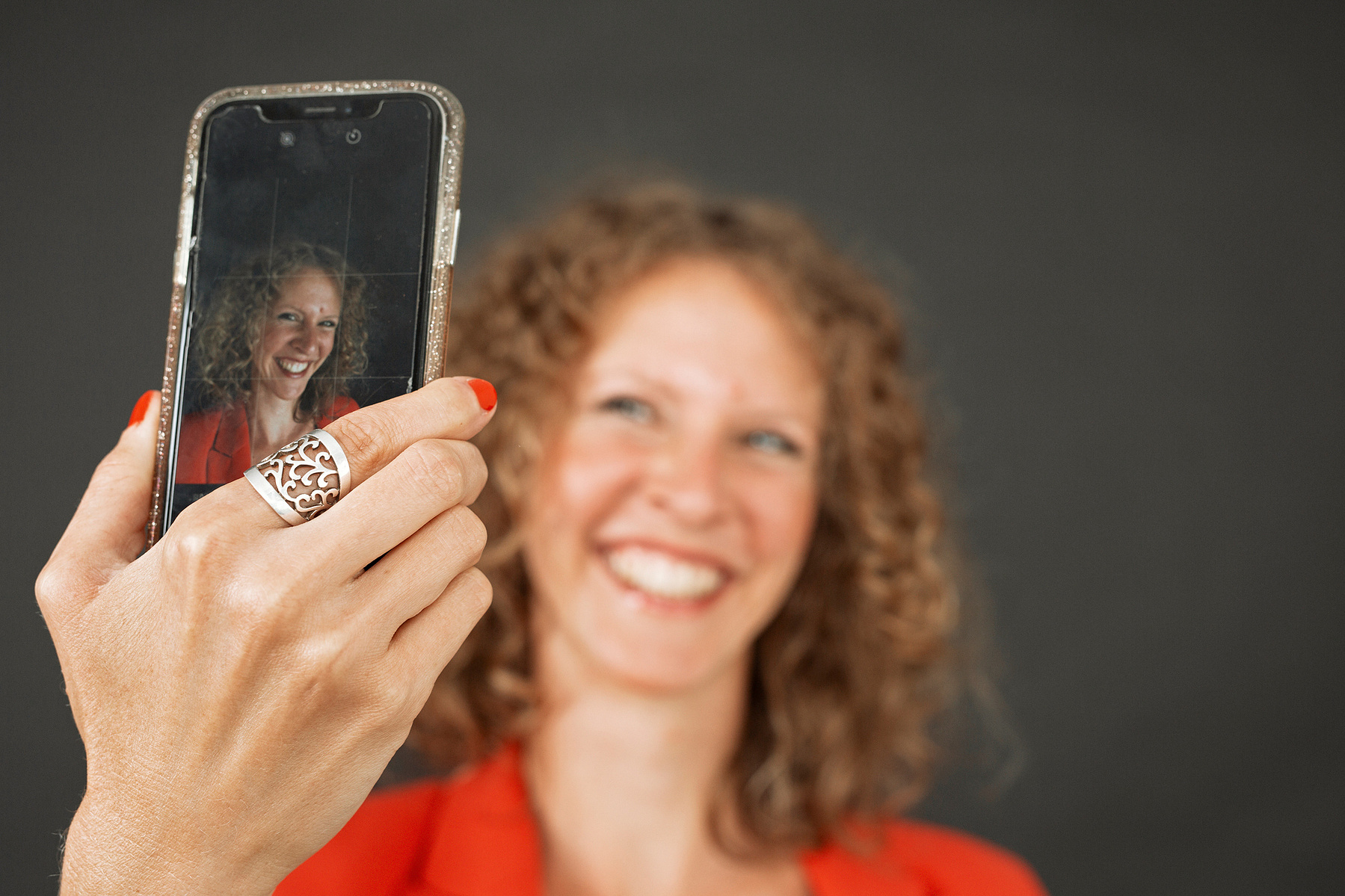 A happy woman with strawberry blonde curls takes a selfie, only the screen of her phone is in focus so we can see the photo she is taking
