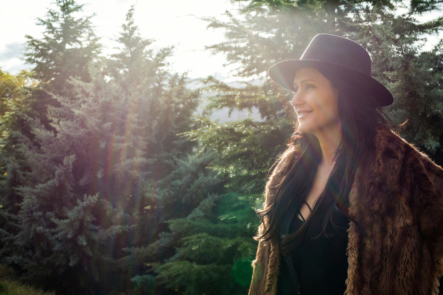 A brunette woman wears a fur jacket and black hat, she is outside and the sun cascades through trees