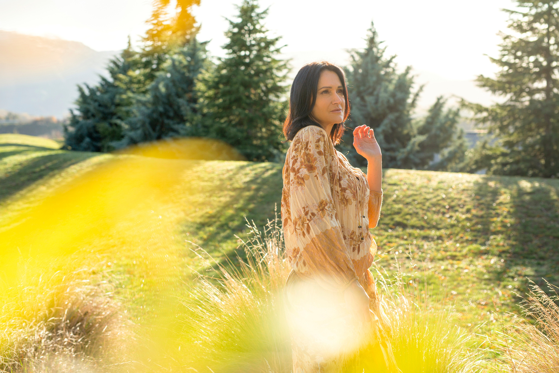 A brunette woman is framed by trees, the mountains of Queenstown visible behind her