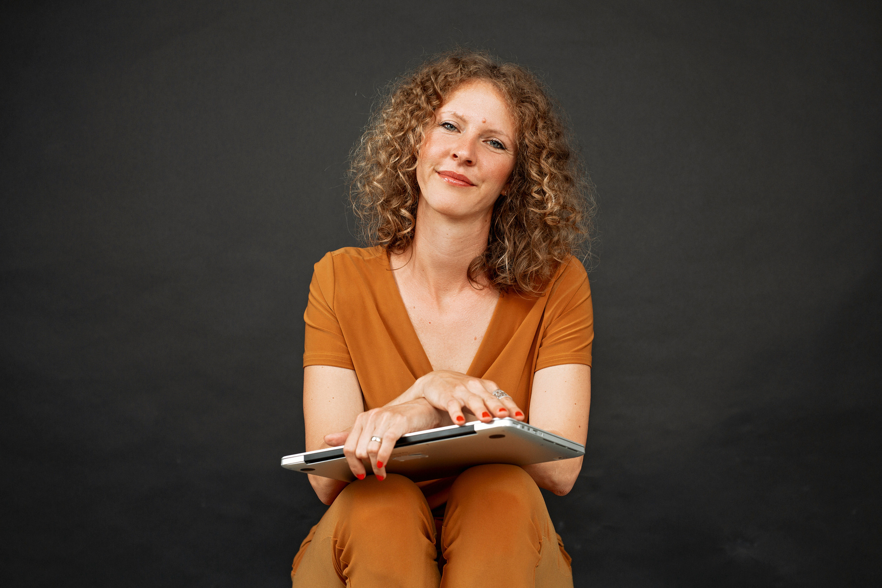 A slim woman sits with her laptop on her knees, a half smile on her lips as she looks at the camera