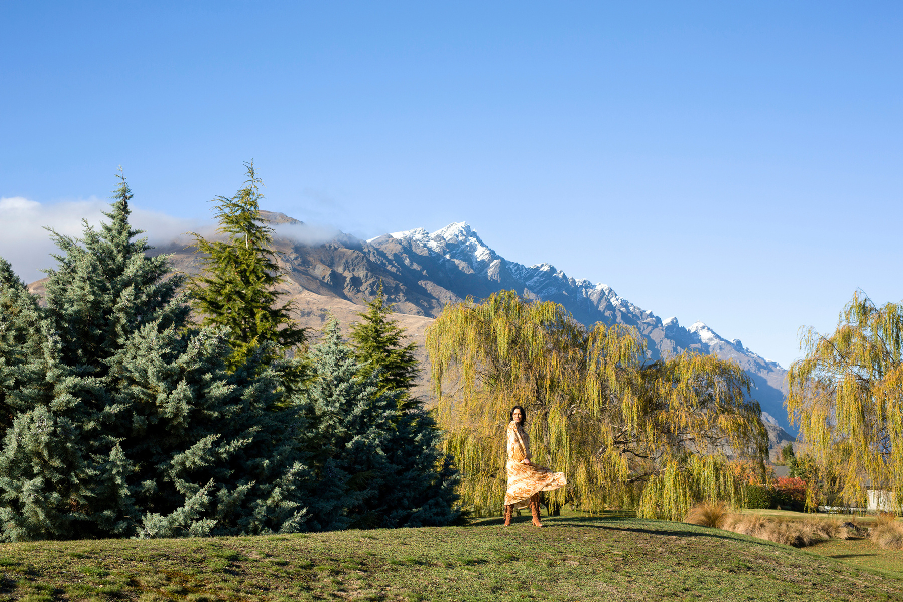 A wide shot of a brunette woman in a floral dress, behind her are the mountains ranges of Queenstown