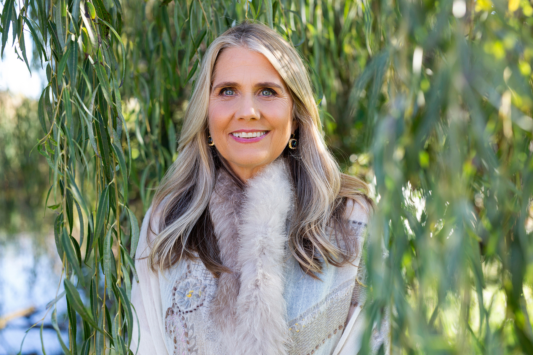 A glamourous older woman stands in front of a willow tree, she is smiling brightly at the camera