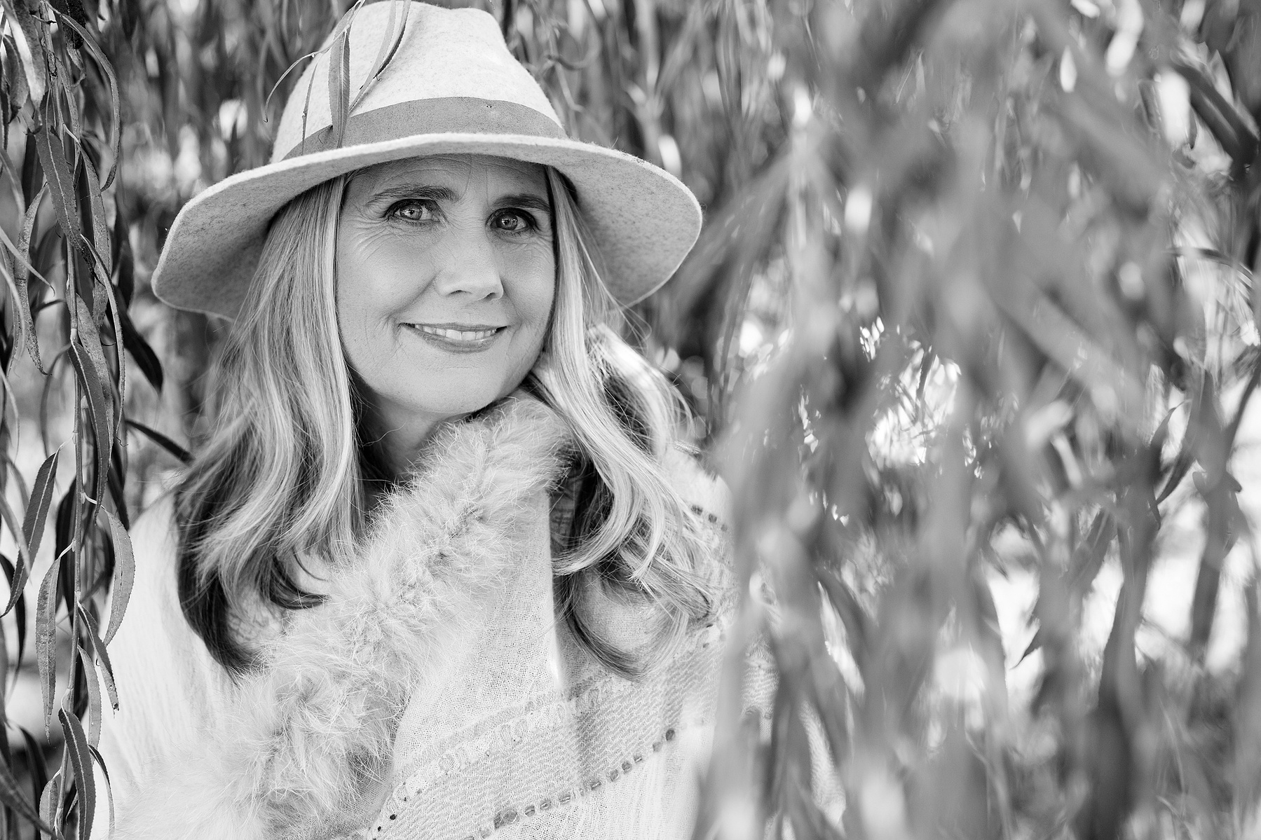 A blonde woman wearing a hat is framed by the branches of a willow tree