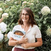 A vibrant brunette woman stands in front of a large plant holding a whole30 cookbook