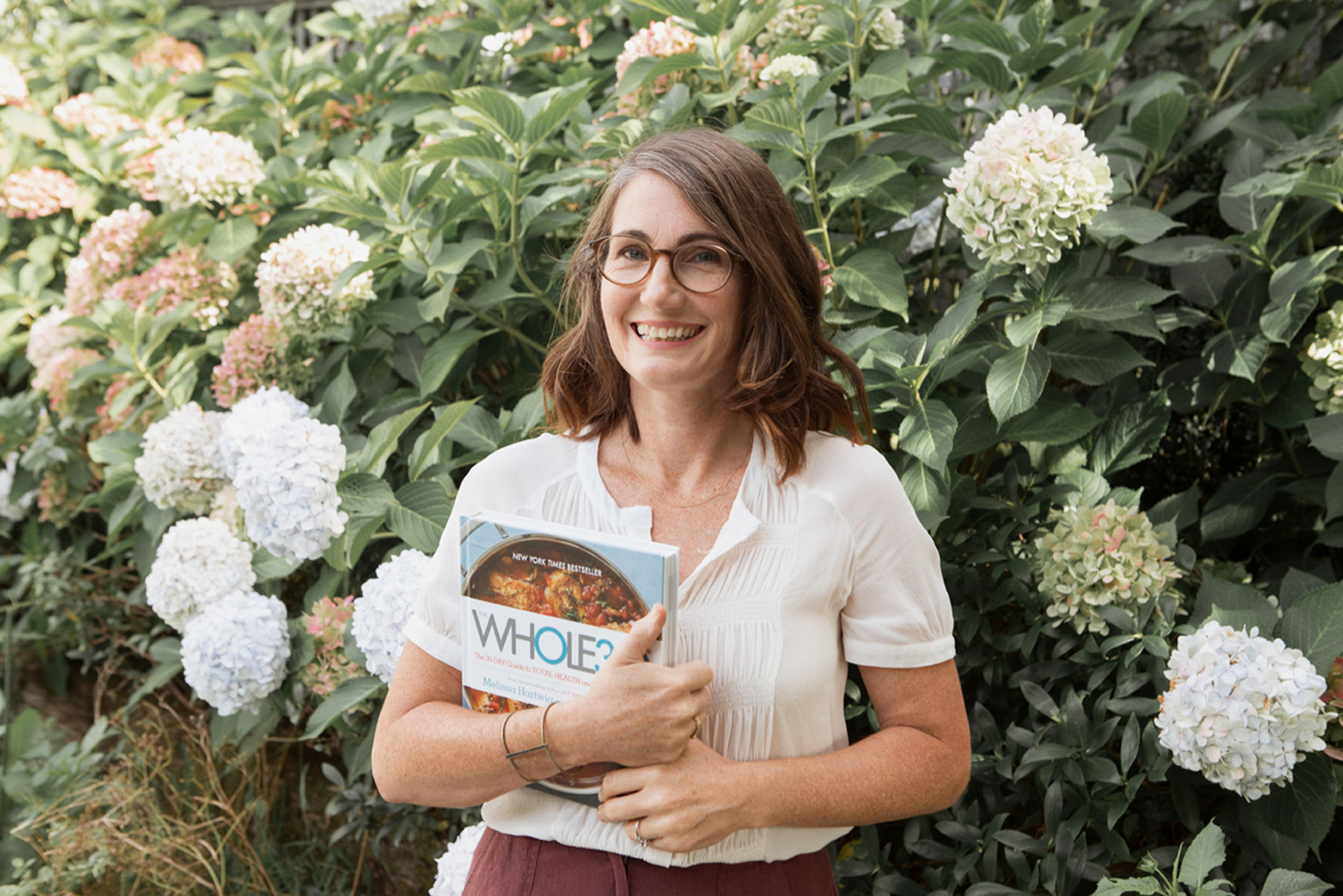 A vibrant brunette woman stands in front of a large plant holding a whole30 cookbook