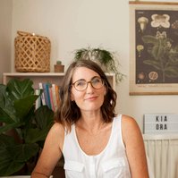 A woman with a brunette bob sits in her home, the smiles enigmatically at the camera