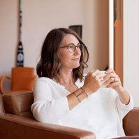 A brunette woman sits in a pink armchair, wearing a white shirt and holding a cup of tea
