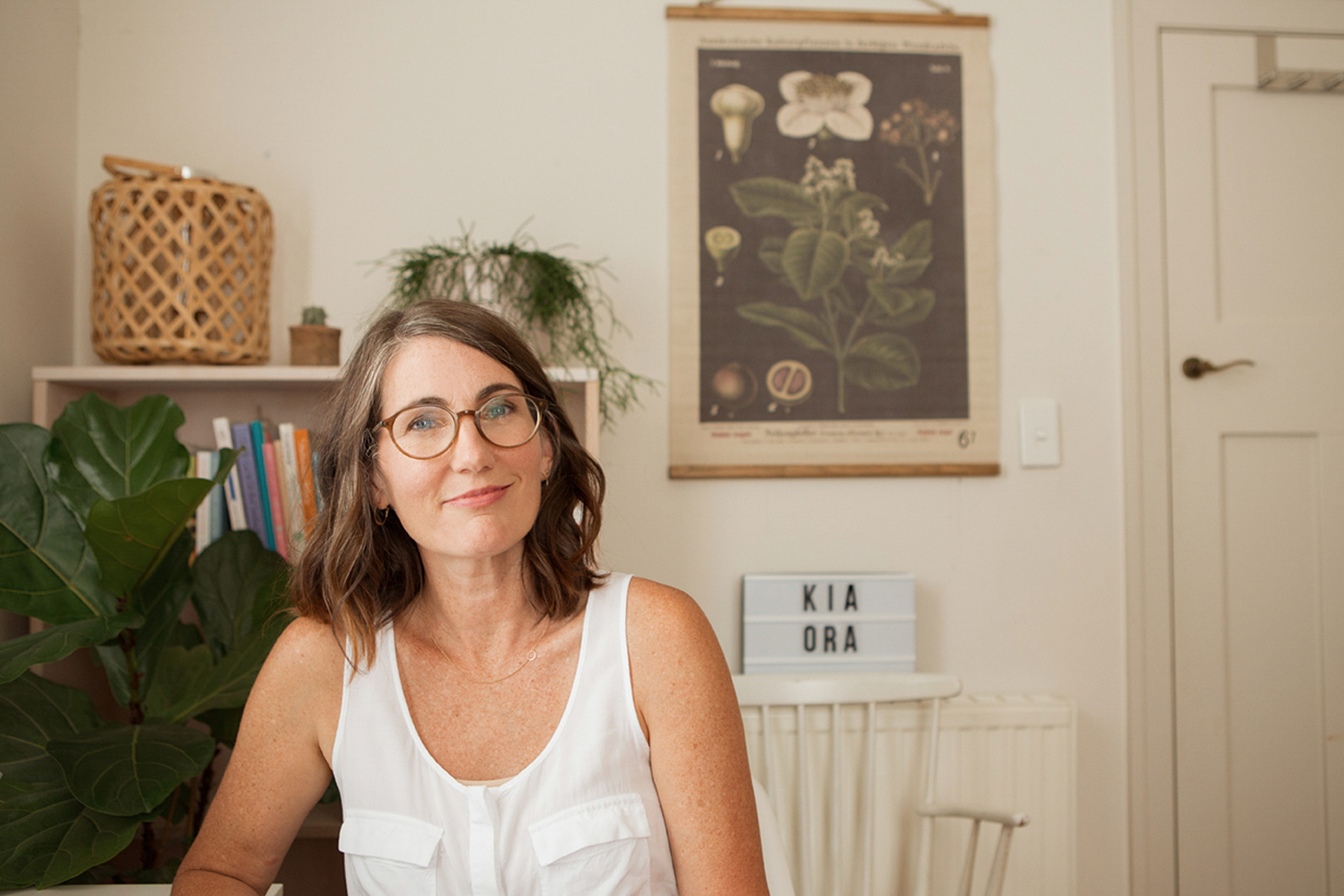 A woman with a brunette bob sits in her home, the smiles enigmatically at the camera
