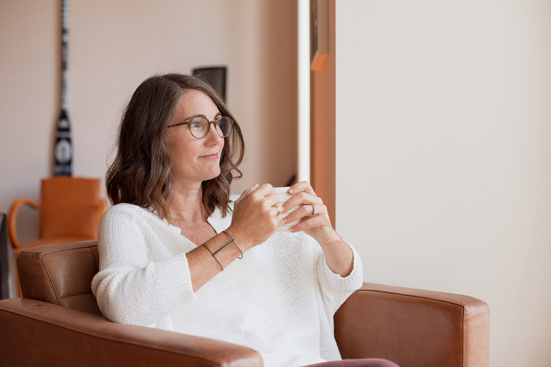 A brunette woman sits in a pink armchair, wearing a white shirt and holding a cup of tea
