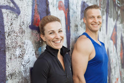 Two personal trainers smile at the camera as they lean against a graffiti covered wall