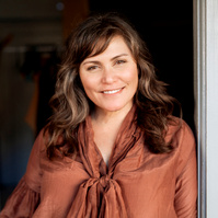 A woman with wavy brown hair leans in a doorway, smiling at the camera
