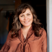 A woman with wavy brown hair leans in a doorway, smiling at the camera