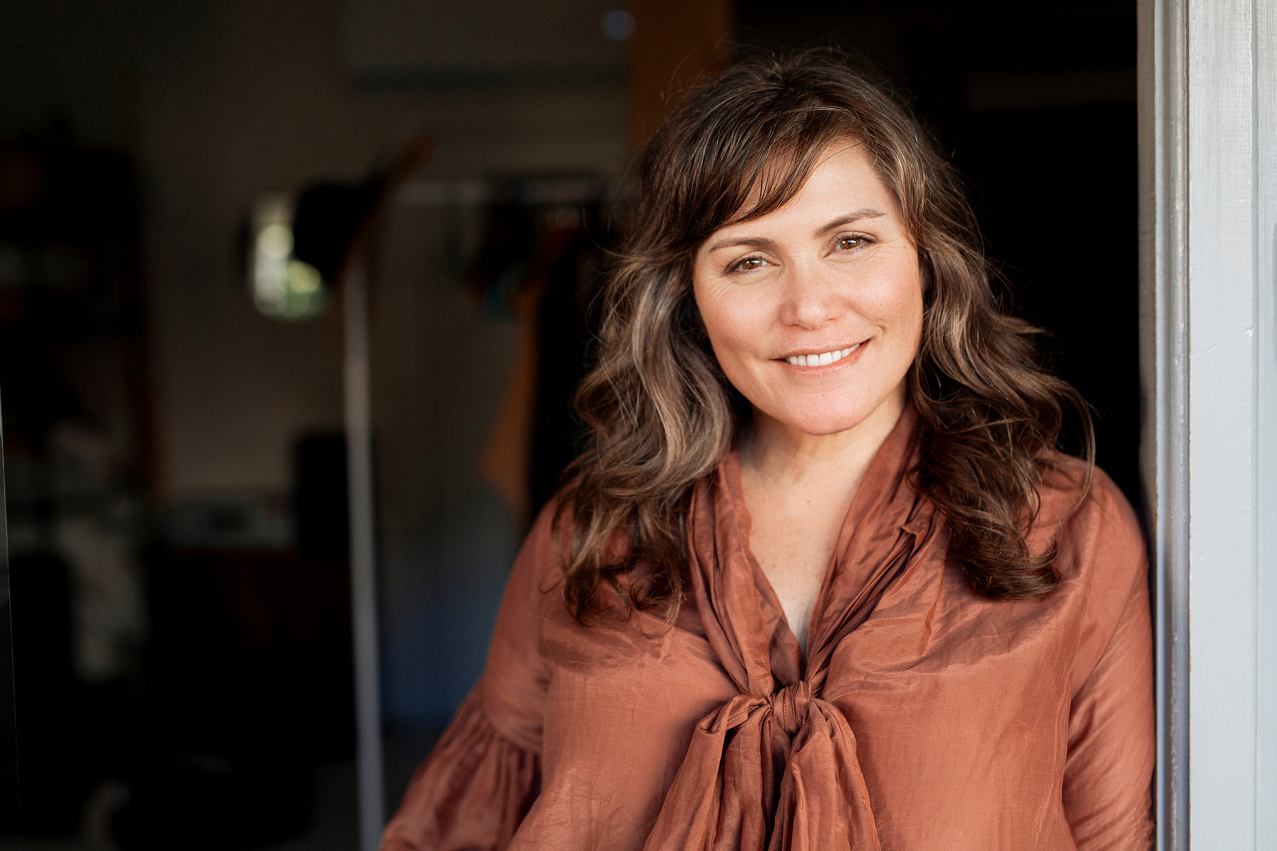 A woman with wavy brown hair leans in a doorway, smiling at the camera