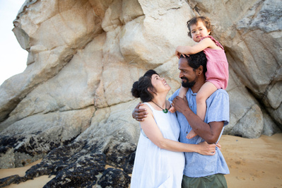 A pregnant woman embraces her husband who is carrying their daughter on his shoulders. They are standing on a sunny beach with large rocks behind them.