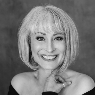 A slim older woman with a blonde bob smiles at the camera. A hand rests on her chest and she wears a dark off the shoulder top. 