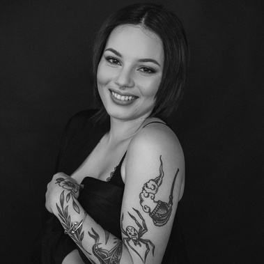 A slim tattooed brunette woman poses in a boudoir style, smiling at the camera and displays her tattoos