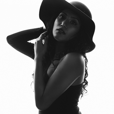A young woman with dark hair stares at the camera, she is wearing a wide-brimmed hat. The background is light and she is dark