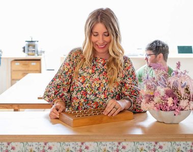 A blonde woman smiles while looking down at a tray of hand made rings