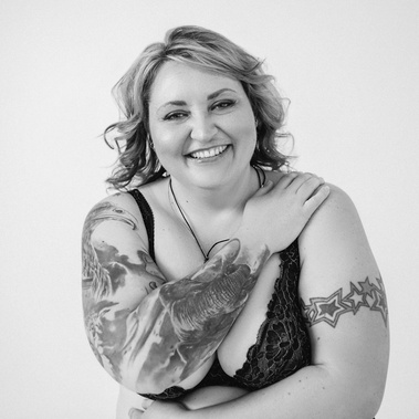 A curvaceous woman with tattoos smiles at the camera, arms covering her chest and stomach
