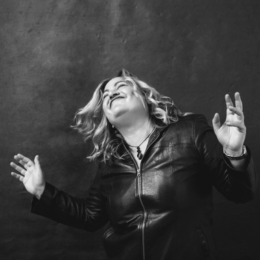 A curvaceous blonde woman appears to be dancing. She is wearing a black leather jacket and her hands are thrown out