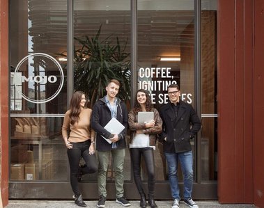 Four people stand outside of the cafe where they work, they look happy