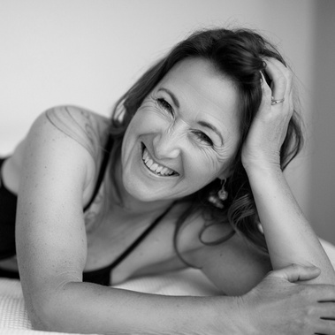 A slim white middle aged woman is lying down leaning her head on a hand, she is smiling at the camera