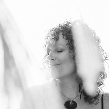 A woman with brown ringlets smiles with her eyes closed, she wears a chunky necklace and is partially obscured by light coloured fabric.