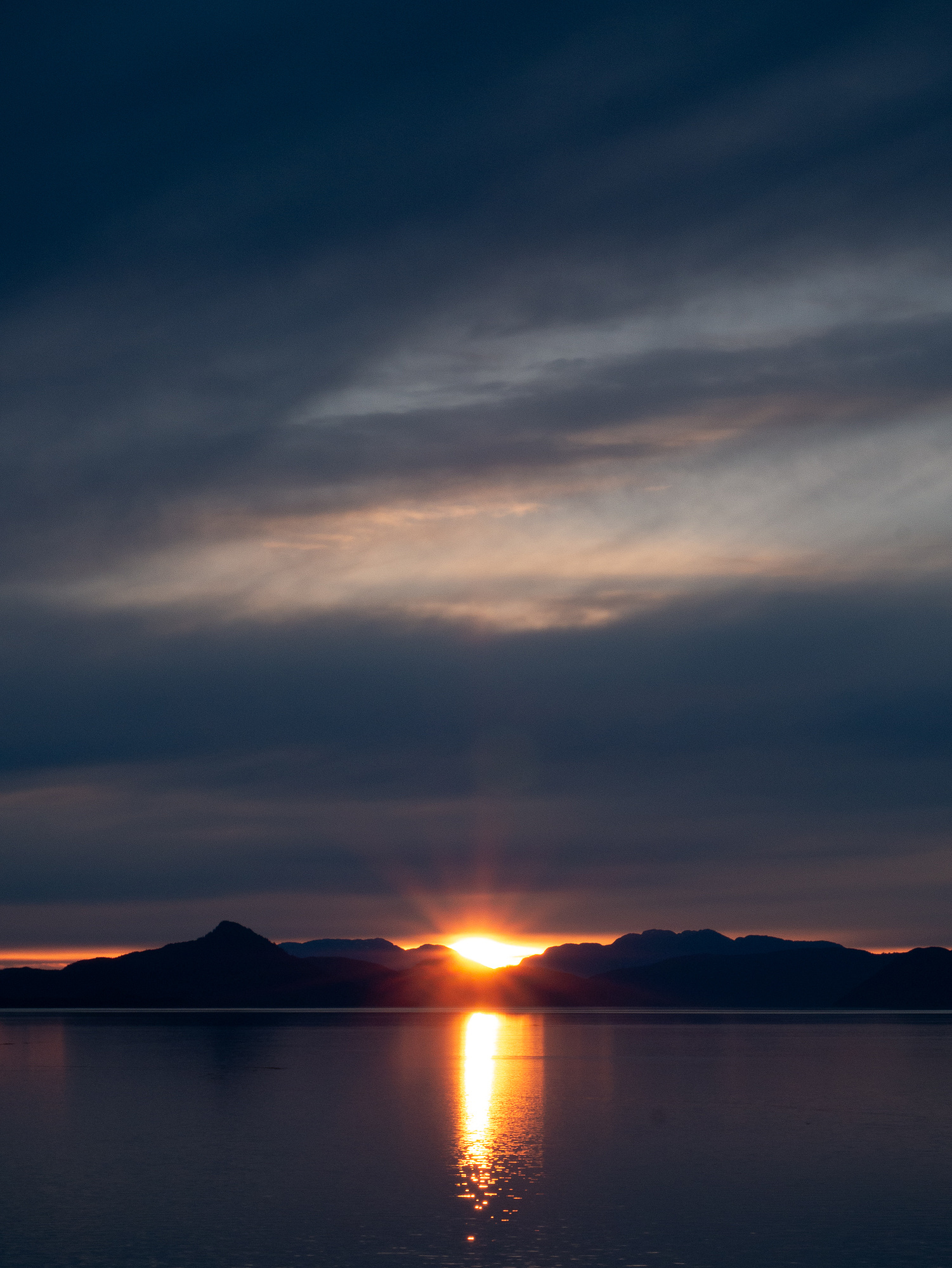 The sun dips behind Alaskan peaks, casting a fiery glow over calm waters, with clouds adding drama to the twilight sky.