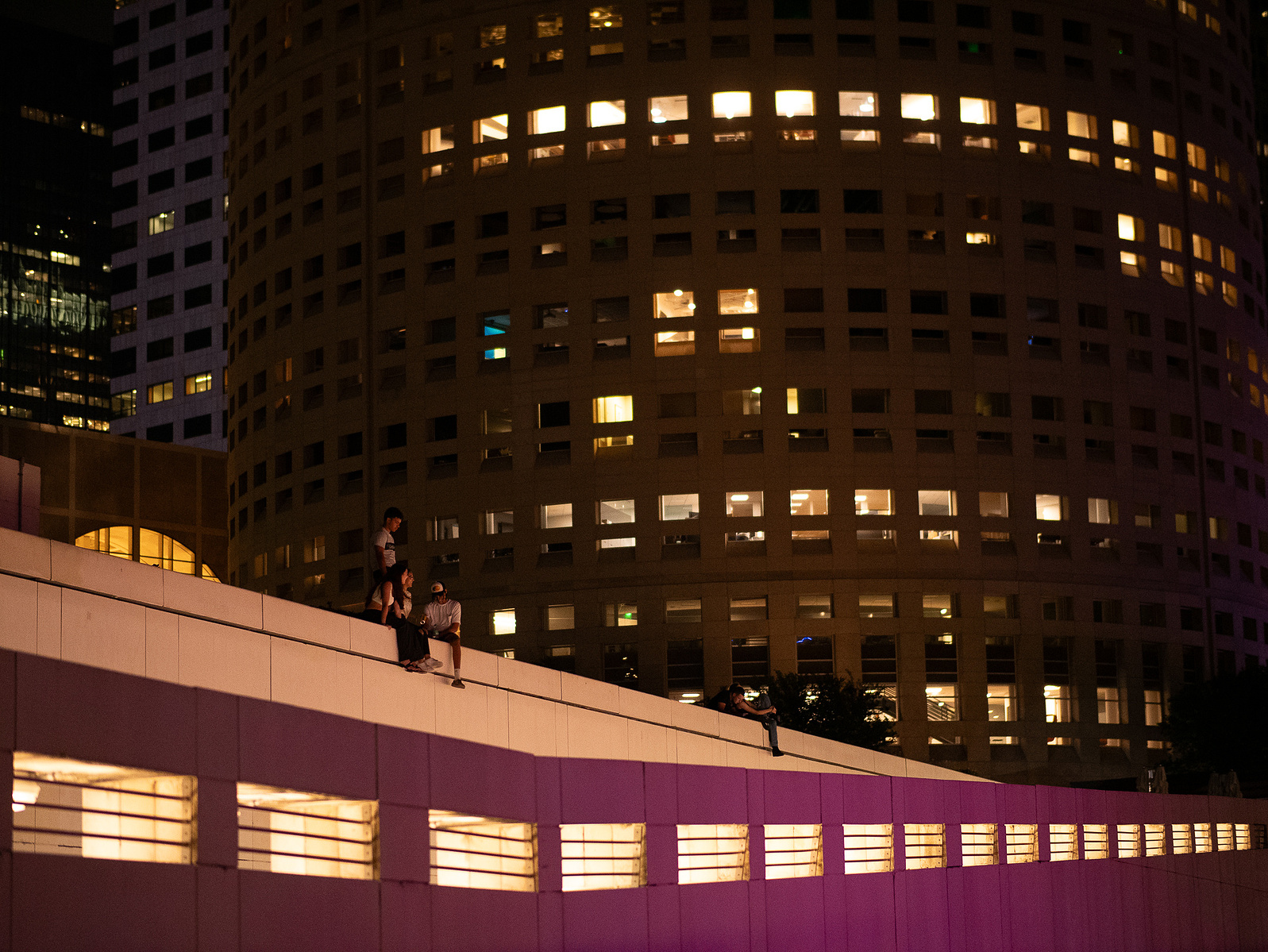 Individuals perched on the brink of land beside Tampa's Sykes building, overlooking the riverwalk and river, under the city lights.