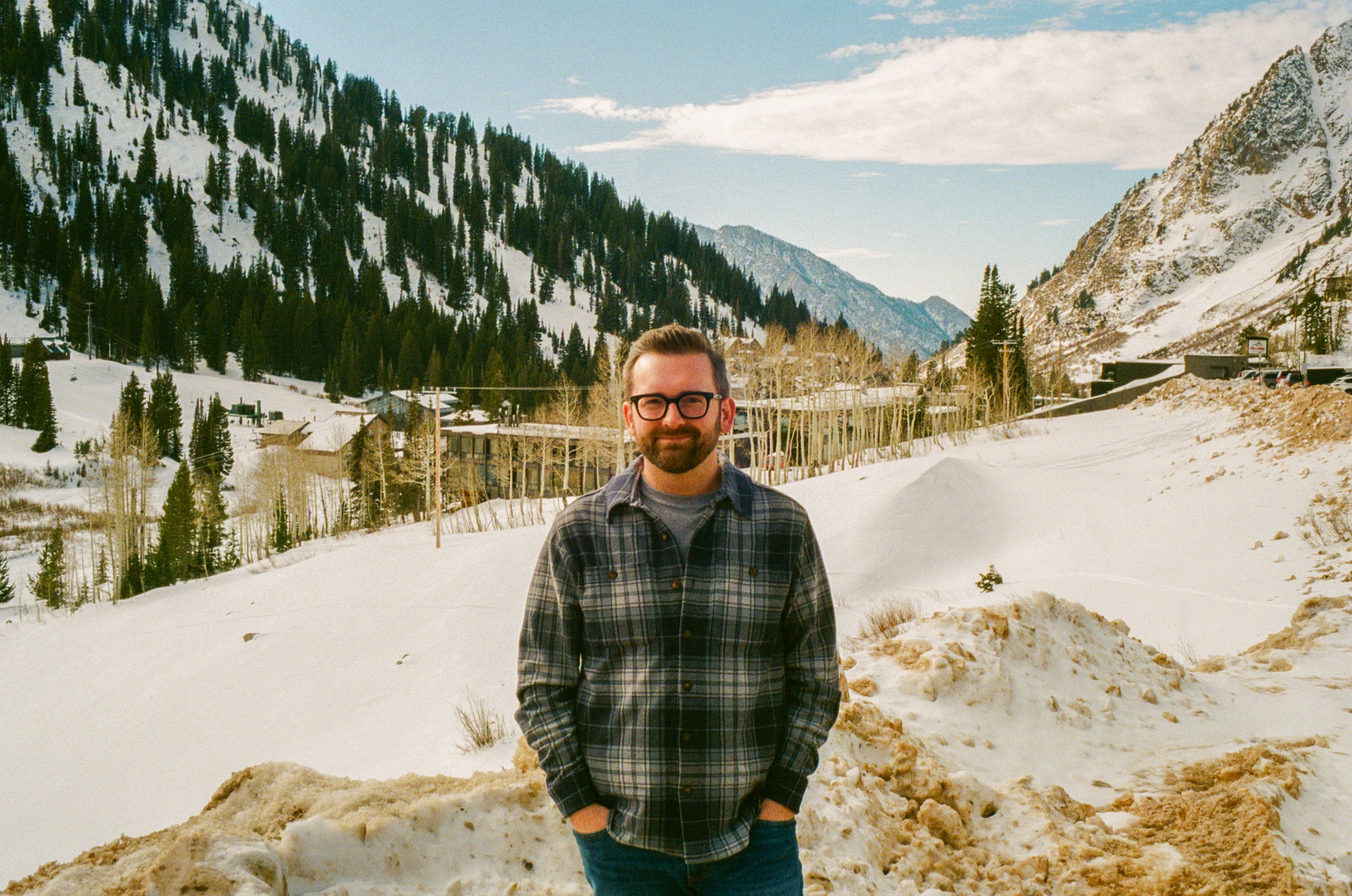 Zack standing in front of skiers, snow drift, and mountains with blue sky
