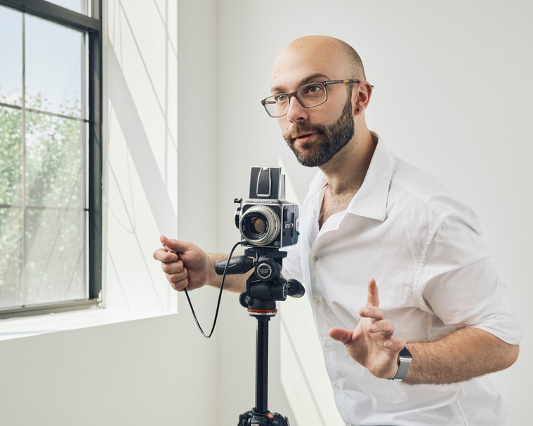 A portrait of a young, white man in a studio. A window illuminates him from the left. In his hand, he holds a shutter release connected to a vintage film camera on a tripod.