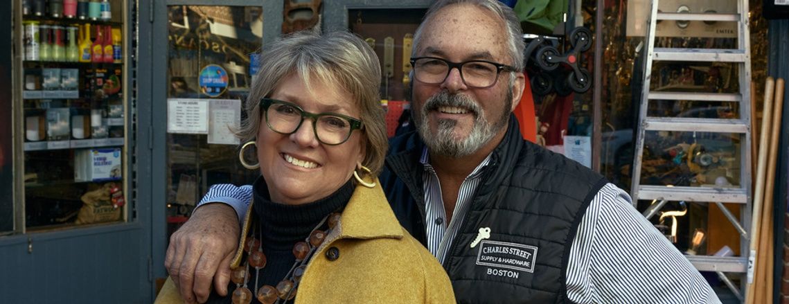 A portrait of a woman in a yellow coat and a man in s striped shirt and vest standing in front of a hardware store.