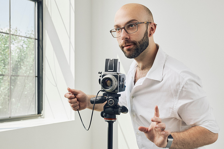 Photo of a young man with a camera on a tripod. The room is bright and he is lit by a window at the left of the image.