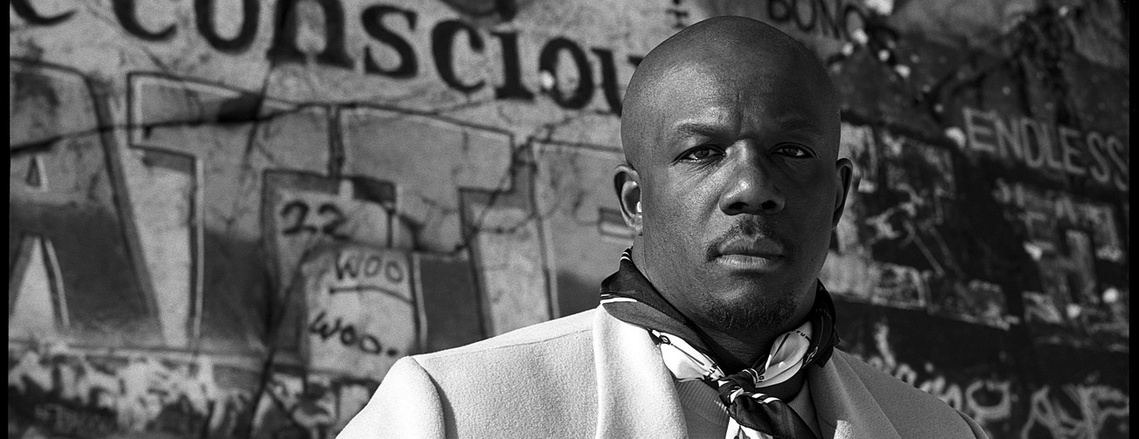 A black and white portrait of a Black man in a white coat and silk scarf. He stands in front of a wall covered in graffiti at the Quincy Quarries in Quincy, MA