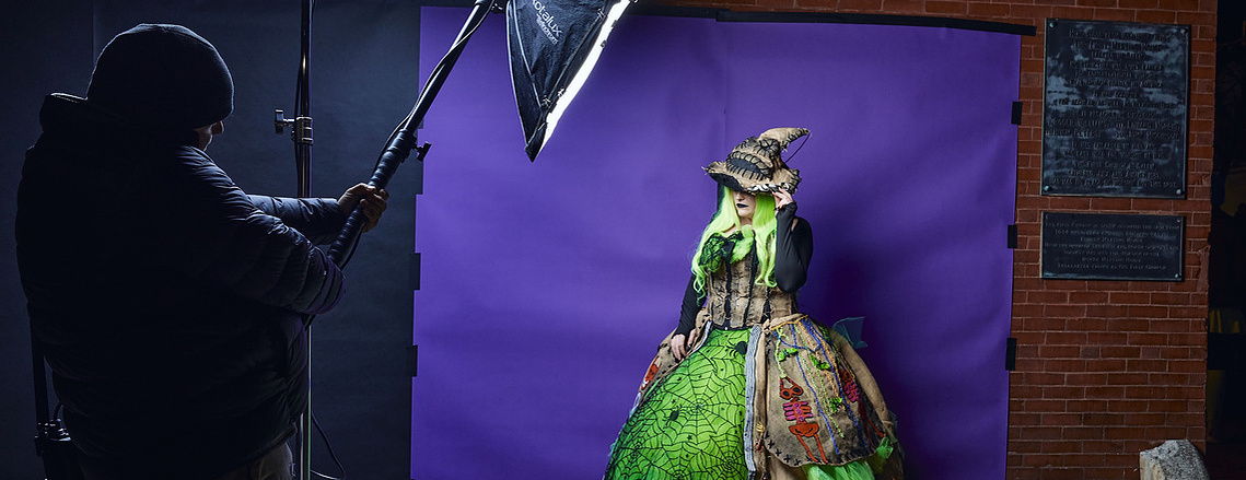 A photo of a woman in an elaborate Halloween costume covered in spiders, wearing a witch hat. She stands in front of a purple backdrop taped to a wall. A man holds a strobe in an umbrella, illuminating her.