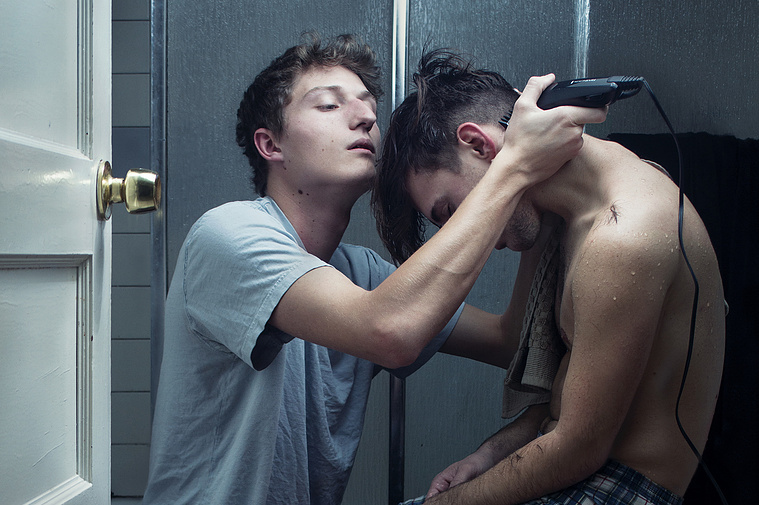 A detail of a portrait with two men in a bathroom. One man uses an electric razor to shave the other man's hair.