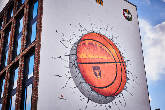 A large building with many windows has a painting of a large basketball on the side. The basketball looks as if it has been thrown into the wall and cracked it
