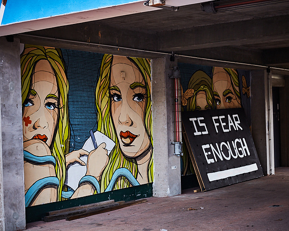 A painting of a blonde woman wearing a blue dress. She is painted twice, both faces facing towards each other. In front of the painted woman is a sign that says 