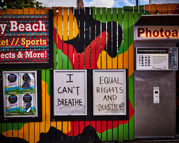 Photo of two empty poster cases on a wooden fence. The fence is painted black, green, red and orange. On one of the poster frames someone has written in capital letters 