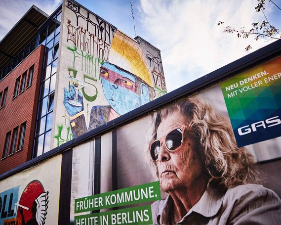 An billboard advert of a senior lady wearing sunglasses below a street artists work of a blue and purple-faced man. The woman in the advert and the street artists cartoon are looking in the same direction, as if they are looking at the same things