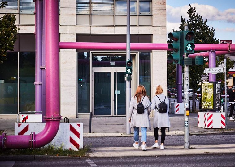 Two women cross the road in Berlin. They both wear long grey coats + small black rucksacks and have the same auburn colour hair. A green Ampelmännchen can be seen on the lights. The shot is framed by large pink pipes going over the road