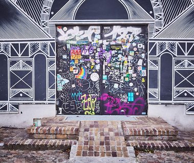 Black and white paint covers the walls of a building, it's painted with triangular shapes, lines and curves. In the centre there are steps leading up to a door covered in stickers and graffiti tags
