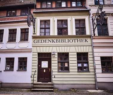 A green building sandwiched between two pink buildings. The green one is a library reading 'Gedenkbibliothek'on the front in large black letters